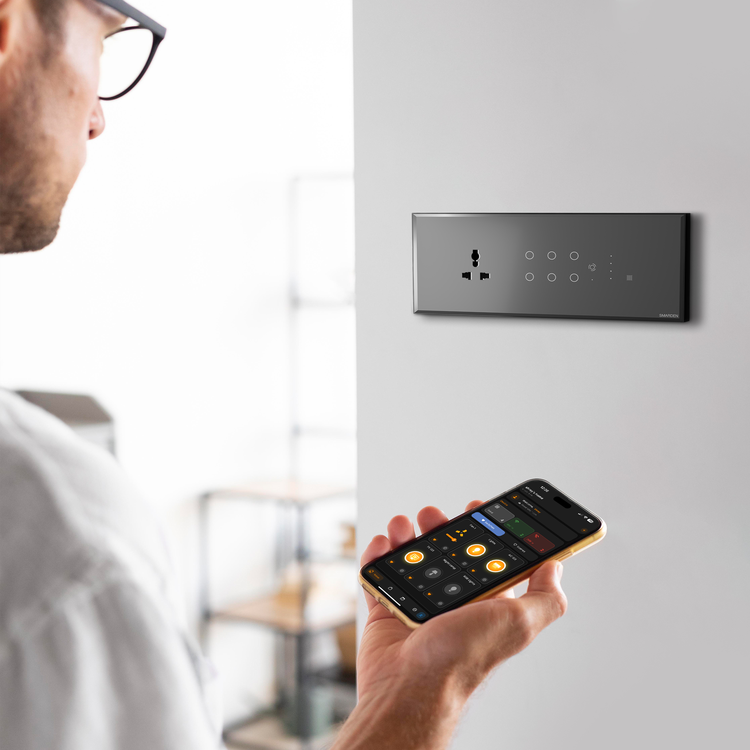 Person using SmarDen mobile app on smartphone to control wall-mounted smart touch switch board